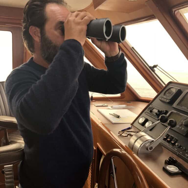 Man at the helm of a powerboat looking out the front window through binoculars.